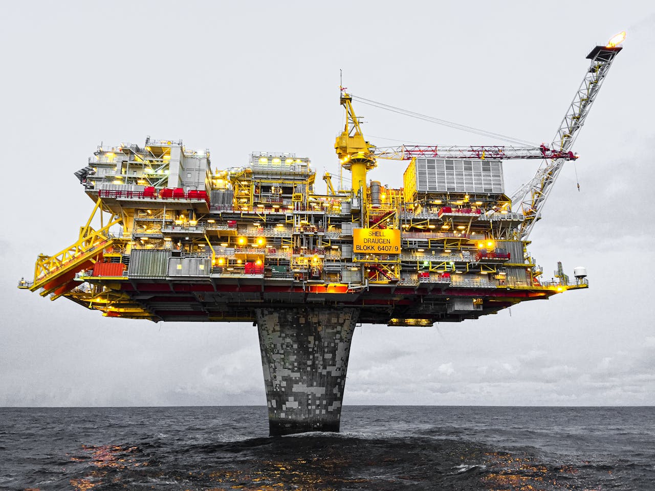 our-services-1 A well-lit offshore oil platform against a cloudy sky in Norway's waters.